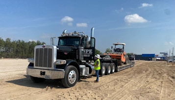 A large black semi-truck with a chrome grille is parked on a sandy construction site. The truck is towing a flatbed trailer carrying an orange heavy-duty construction vehicle. A person wearing a white hard hat and yellow safety vest stands next to the truck. The site is surrounded by dirt and some construction equipment, with trees and blue sky in the background.