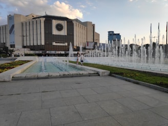A large modern building with a distinctive emblem on its facade stands behind a series of fountains. The structure is surrounded by a spacious paved area with neatly trimmed flowerbeds to the side. The sky is partly cloudy, and in the foreground, a couple of people walk along the path next to the fountains, enjoying the scenery.