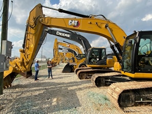 Several large construction excavators are lined up in a row on a gravel surface. Two people wearing hard hats and casual clothing are standing next to the machinery, appearing to be discussing. The sky is partly cloudy, providing a bright background to the scene.