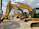 Several large construction excavators are lined up in a row on a gravel surface. Two people wearing hard hats and casual clothing are standing next to the machinery, appearing to be discussing. The sky is partly cloudy, providing a bright background to the scene.