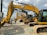 Several large construction excavators are lined up in a row on a gravel surface. Two people wearing hard hats and casual clothing are standing next to the machinery, appearing to be discussing. The sky is partly cloudy, providing a bright background to the scene.
