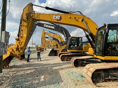 Several large construction excavators are lined up in a row on a gravel surface. Two people wearing hard hats and casual clothing are standing next to the machinery, appearing to be discussing. The sky is partly cloudy, providing a bright background to the scene.