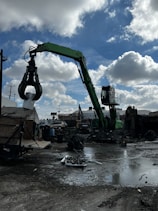 A green industrial crane with a large claw attachment is operating in a scrapyard. The ground is wet and muddy with scattered metal debris. A partly cloudy sky is seen overhead. Surrounding the crane are various industrial structures, including buildings and fences, with some vehicles in the background.
