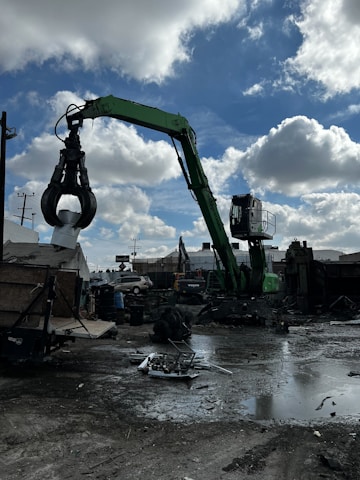 A green industrial crane with a large claw attachment is operating in a scrapyard. The ground is wet and muddy with scattered metal debris. A partly cloudy sky is seen overhead. Surrounding the crane are various industrial structures, including buildings and fences, with some vehicles in the background.