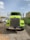 A large, bright yellow-green truck is parked on a gravel surface in an industrial area. The truck has a prominent chrome front grille and two side mirrors extending outwards. There are several parked cars and a green dumpster in the background, with some buildings and power lines visible under a clear, slightly cloudy sky.