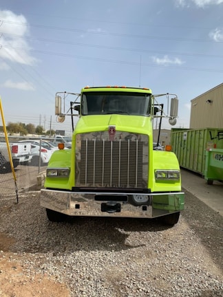 A large, bright yellow-green truck is parked on a gravel surface in an industrial area. The truck has a prominent chrome front grille and two side mirrors extending outwards. There are several parked cars and a green dumpster in the background, with some buildings and power lines visible under a clear, slightly cloudy sky.