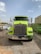 A large, bright yellow-green truck is parked on a gravel surface in an industrial area. The truck has a prominent chrome front grille and two side mirrors extending outwards. There are several parked cars and a green dumpster in the background, with some buildings and power lines visible under a clear, slightly cloudy sky.