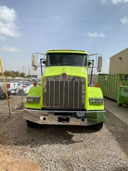 A large, bright yellow-green truck is parked on a gravel surface in an industrial area. The truck has a prominent chrome front grille and two side mirrors extending outwards. There are several parked cars and a green dumpster in the background, with some buildings and power lines visible under a clear, slightly cloudy sky.