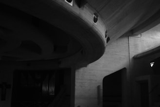 The image depicts an interior space with a modern architectural style, featuring curved concrete structures and a dark, minimalistic design. The atmosphere is dimly lit, highlighting the smooth textures and unique shapes of the architecture. A set of organ pipes is visible in the background, adding to the ambiance.