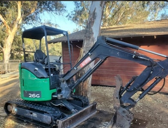 A small excavator with a green and black body is parked outdoors near a wooden building. It has caterpillar tracks and a large mechanical arm with a claw attachment. The surroundings include trees and a fence in the background.