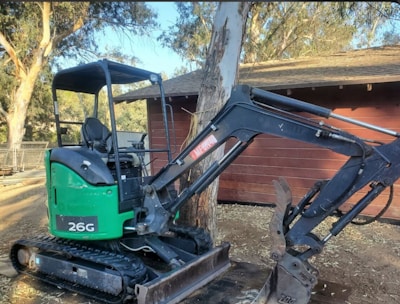 A small excavator with a green and black body is parked outdoors near a wooden building. It has caterpillar tracks and a large mechanical arm with a claw attachment. The surroundings include trees and a fence in the background.