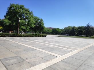 A spacious, paved public square surrounded by lush, green trees under a clear blue sky. The area is open with a few people walking in the distance, and there are tall lampposts. Manicured hedges divide the square from the surrounding greenery which includes tall trees.