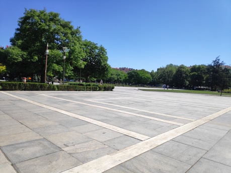 A spacious, paved public square surrounded by lush, green trees under a clear blue sky. The area is open with a few people walking in the distance, and there are tall lampposts. Manicured hedges divide the square from the surrounding greenery which includes tall trees.