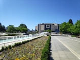 A large, modern building stands prominently in the background, surrounded by lush green trees and plants. In the foreground, colorful flower beds and well-maintained grassy areas are visible, with a series of water fountains lined up parallel to a paved walkway. The sky is clear and blue, adding to the serene atmosphere.