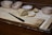 A loaf of bread is partially sliced on a wooden cutting board, surrounded by bread crumbs. A serrated knife with a black handle is placed nearby. Behind the cutting board, four empty wicker baskets are aligned in a row.