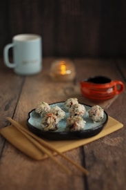 A set of rice balls garnished with vegetables is placed on a speckled blue plate. The food is arranged on a wooden serving board with a pair of chopsticks nearby. In the background, there is a light blue mug, a small red sauce container, and a lit candle in a glass holder.