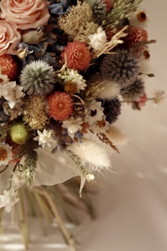 A detailed arrangement of dried and fresh flowers featuring various colors and textures. The bouquet includes soft pink roses, round orange and red dried flowers, blue thistles, bright green buds, and cream-colored fluffy accents. Dried moss and small white flowers add to the rustic and artistic composition.