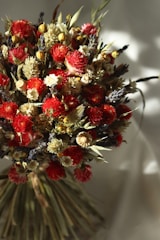 A vibrant bouquet of dried flowers featuring a mix of red, yellow, and cream blossoms. The arrangement includes various textures and shapes, with some flowers appearing fluffy and others tightly packed. Stems and leaves in earthy tones provide contrast. The lighting creates soft shadows, adding depth and a natural feel.