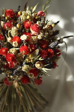 A vibrant bouquet of dried flowers featuring a mix of red, yellow, and cream blossoms. The arrangement includes various textures and shapes, with some flowers appearing fluffy and others tightly packed. Stems and leaves in earthy tones provide contrast. The lighting creates soft shadows, adding depth and a natural feel.