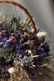A detailed arrangement of dried flowers and grasses in a woven basket, including purple and pink blooms along with various textured herbs and stems.