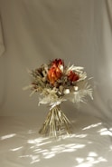 A bouquet of dry flowers with red and orange protea flowers, surrounded by various dried foliage and twigs arranged in a rustic style. The setting is minimalist with a neutral background, casting soft shadows that create an artistic effect.
