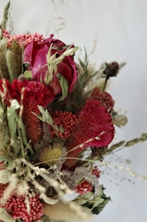 A vibrant and artistic arrangement of flowers featuring deep red and pink blossoms interspersed with dried foliage and grasses. The mix of textures and colors creates a visually striking bouquet.
