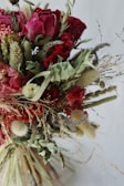 A vibrant bouquet composed of dried flowers, featuring red roses with curled petals, intermingled with various types of grasses and foliage. Soft textures and earthy colors contribute to the bouquet's rustic charm. The arrangement includes fluffy seed pods and curly green leaves that offer a natural, organic feel.