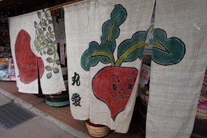 Three fabric panels are hanging outside a shop. Each panel features artwork of radishes with distinct styles and expressions. The radishes are predominantly red with green leaves, and there are Japanese characters printed on the fabric. Various posters and items are visible in the background.