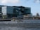 A modern office building with reflective glass windows sits alongside a body of water. The architecture features angular and geometric designs. A small boat is visible on the water in front of the building. The sky is partly cloudy, and there are some bare tree branches in the upper corners of the image.