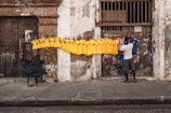 A street vendor is arranging a row of brightly colored yellow and blue soccer jerseys hanging on a line in front of a weathered, old building. The wall is contrastive, with textures of peeling paint and worn surfaces. The vendor is casually dressed and is standing on a cracked sidewalk next to a plastic chair with a dark cover.