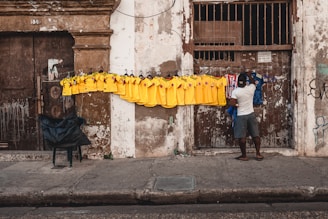A street vendor is arranging a row of brightly colored yellow and blue soccer jerseys hanging on a line in front of a weathered, old building. The wall is contrastive, with textures of peeling paint and worn surfaces. The vendor is casually dressed and is standing on a cracked sidewalk next to a plastic chair with a dark cover.