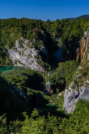 A picturesque landscape featuring lush green forests and rugged rock formations. A series of waterfalls cascade into a serene turquoise lake surrounded by vibrant greenery under a clear blue sky.