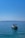 A small motorboat with a blue canopy floats on calm, clear blue water with a distant view of an island or landmass on the horizon beneath a bright blue sky.
