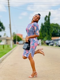 A person poses joyfully on a paved pathway, wearing a colorful dress with floral patterns and light brown heels. The background features green trees and parked cars under a bright blue sky.