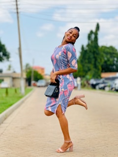 A person poses joyfully on a paved pathway, wearing a colorful dress with floral patterns and light brown heels. The background features green trees and parked cars under a bright blue sky.