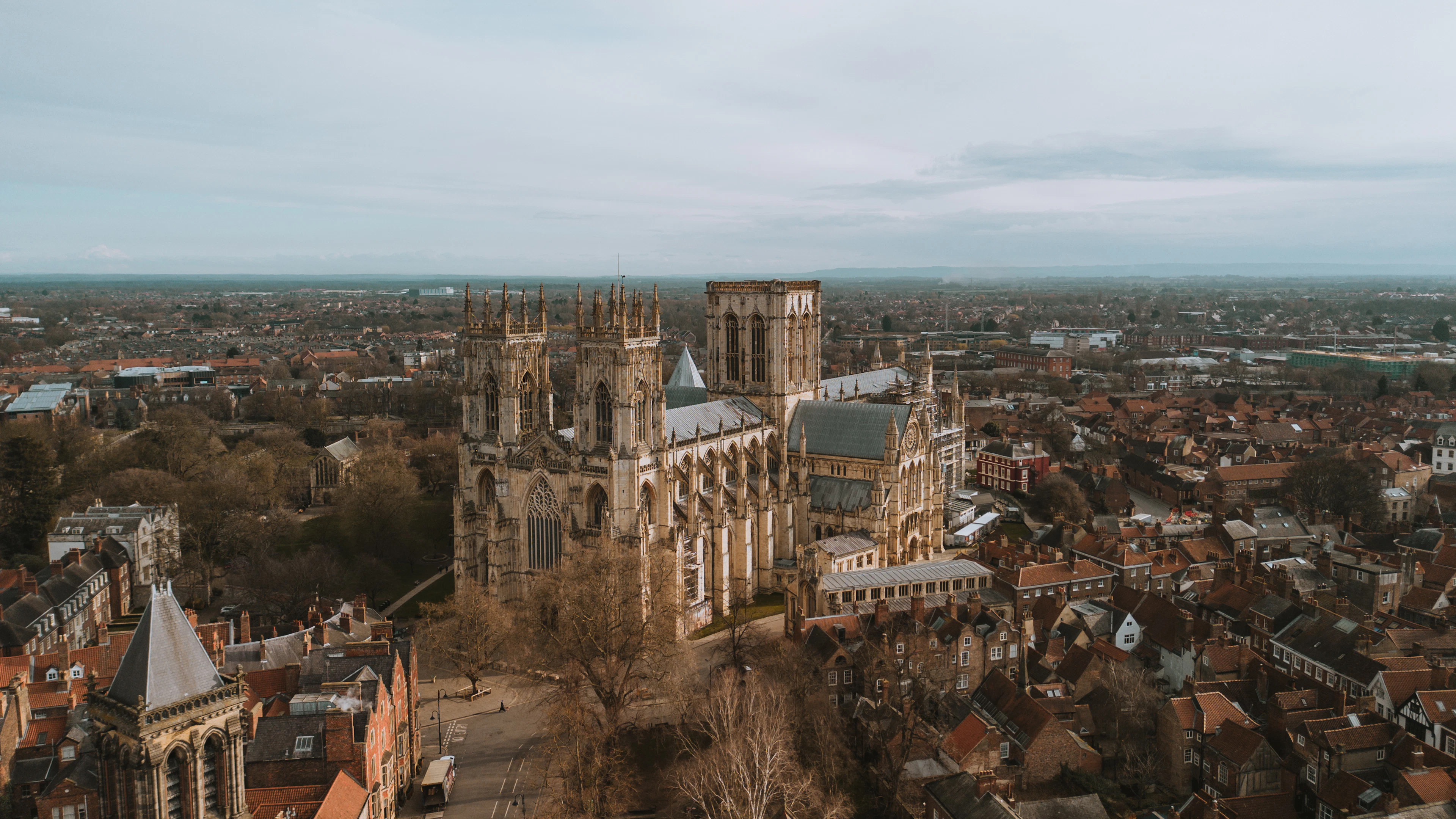 York Minster seen from the air, its twin west towers and long nave rising out of the red-tiled rooftops of the walled city, with the Vale of York stretching flat to the horizon.