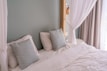 A neatly made bed with white linens and two decorative pillows featuring a geometric pattern in blue tones. The bed is framed by a light green wall and partly enclosed by sheer white curtains. Natural light filters in through a nearby window with beige drapes.