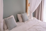 A neatly made bed with white linens and two decorative pillows featuring a geometric pattern in blue tones. The bed is framed by a light green wall and partly enclosed by sheer white curtains. Natural light filters in through a nearby window with beige drapes.