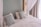 A neatly made bed with white linens and two decorative pillows featuring a geometric pattern in blue tones. The bed is framed by a light green wall and partly enclosed by sheer white curtains. Natural light filters in through a nearby window with beige drapes.