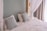 A neatly made bed with white linens and two decorative pillows featuring a geometric pattern in blue tones. The bed is framed by a light green wall and partly enclosed by sheer white curtains. Natural light filters in through a nearby window with beige drapes.