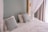 A neatly made bed with white linens and two decorative pillows featuring a geometric pattern in blue tones. The bed is framed by a light green wall and partly enclosed by sheer white curtains. Natural light filters in through a nearby window with beige drapes.