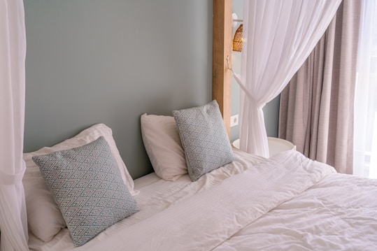 A neatly made bed with white linens and two decorative pillows featuring a geometric pattern in blue tones. The bed is framed by a light green wall and partly enclosed by sheer white curtains. Natural light filters in through a nearby window with beige drapes.