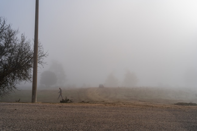 Still from a black-and-white video showing a lone figure walking through a foggy street at dawn.