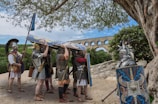 A group of individuals dressed as Roman soldiers gather under a tree. They hold shields adorned with intricate designs and wear armor typical of ancient Roman times. In the background, there is a large stone aqueduct against a clear blue sky, and the surrounding landscape is lush and green.