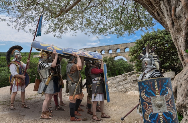 A group of individuals dressed as Roman soldiers gather under a tree. They hold shields adorned with intricate designs and wear armor typical of ancient Roman times. In the background, there is a large stone aqueduct against a clear blue sky, and the surrounding landscape is lush and green.