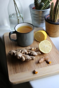 A wooden cutting board holds a mug filled with yellow liquid, fresh ginger roots, a sliced lemon, and turmeric. In the background, there is a glass bottle with water and two potted cacti.