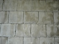 A pattern of rectangular concrete tiles arranged in a staggered layout, creating a textured surface. The tiles have a rough, slightly uneven surface with visible imperfections and a muted greenish-gray color.