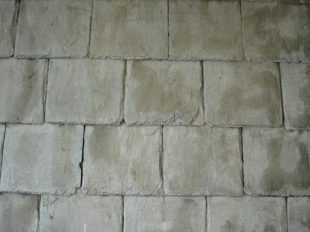 A pattern of rectangular concrete tiles arranged in a staggered layout, creating a textured surface. The tiles have a rough, slightly uneven surface with visible imperfections and a muted greenish-gray color.