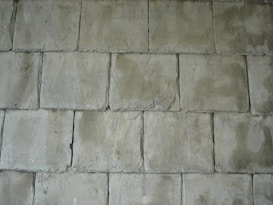 A pattern of rectangular concrete tiles arranged in a staggered layout, creating a textured surface. The tiles have a rough, slightly uneven surface with visible imperfections and a muted greenish-gray color.