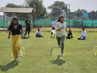 A group of people is enjoying an outdoor activity on a grassy field. Two women in the foreground are participating in a hurdle race, with small yellow hurdles placed in their path. Several other individuals are seated or standing in the background, observing the race. The setting includes trees and a gazebo, fenced off with green mesh.
